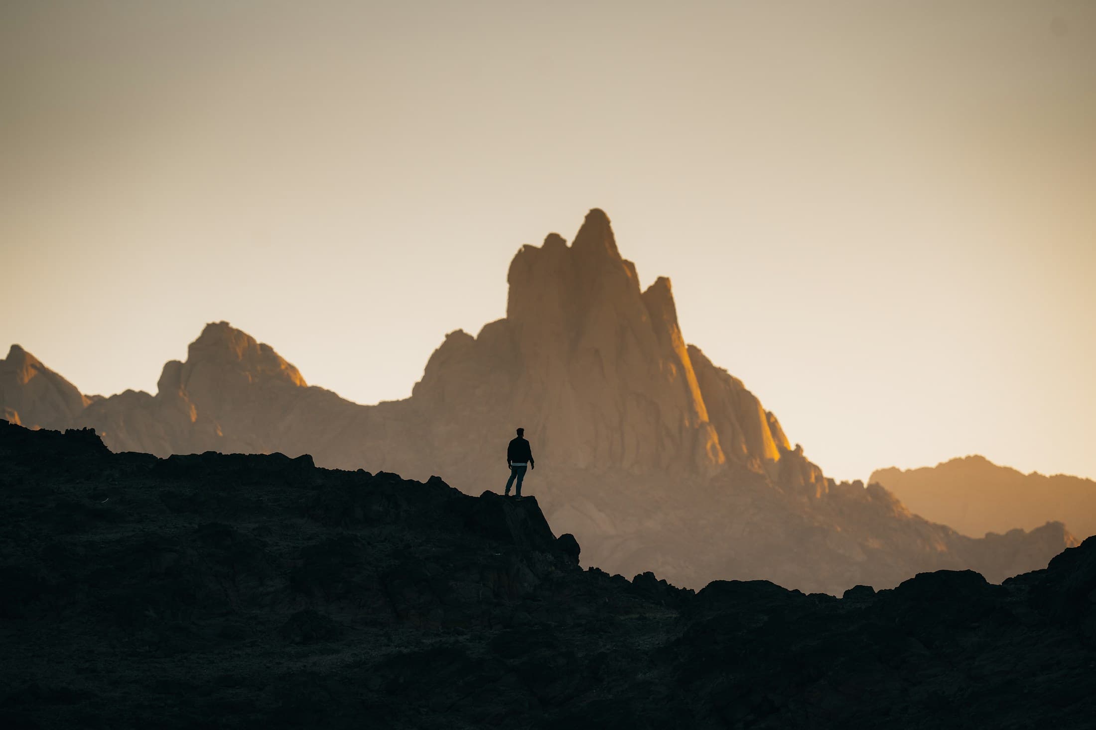 A lone figure on a rocky ridge facing a sunlit mountain peak at golden hour